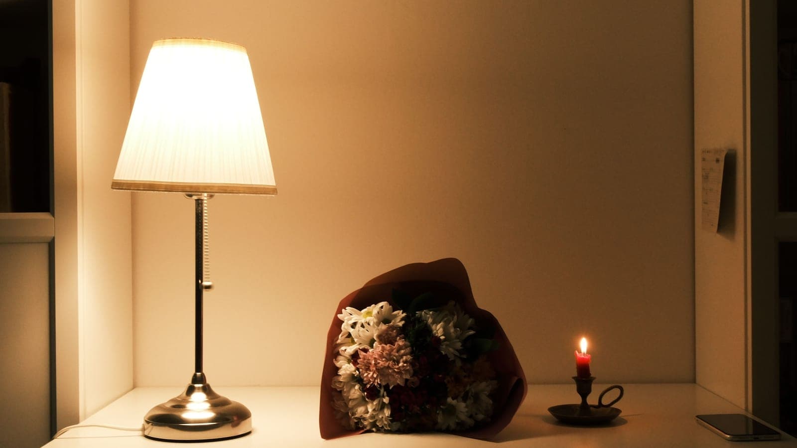 A lit table lamp with a white pleated shade beside flowers and a candle on a shelf